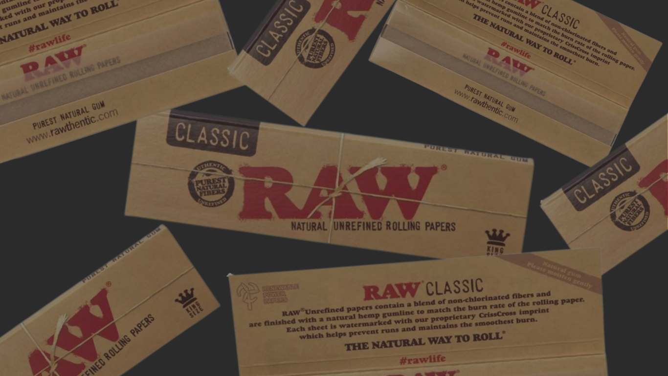RAW Classic King Size Longpapes Banner: mehrere braune RAW-Classic-Packungen und geöffnete Hefte auf dunkelgrauem Hintergrund, mittig eine große RAW-Classic-King-Size-Packung, drumherum weitere Packungen in verschiedenen Winkeln.